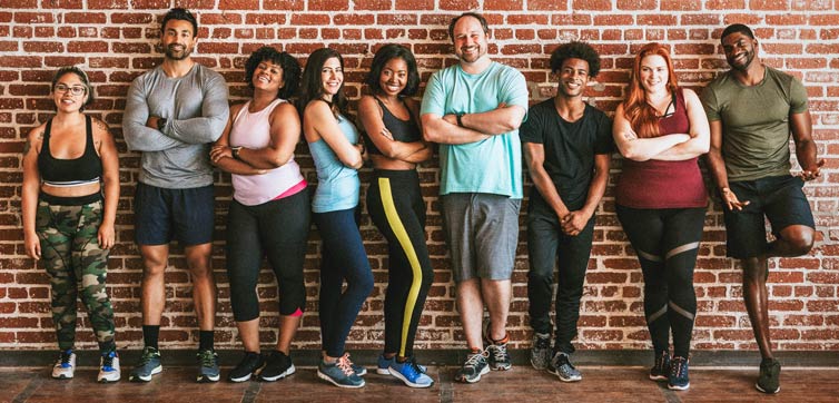 Mixed group (age, race, sex) of smiling people in workout clothes leaning against a brick wall.