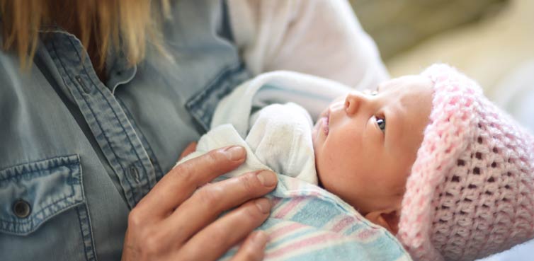 Woman holding infant wearing a pink cap wrapped in blanket.