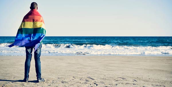 Person standing with back to viewer on the shore with a rainbow flag draped across their shoulders.