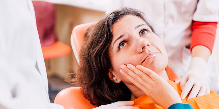 Woman in a dentist’s chair holding her right hand against her jaw.