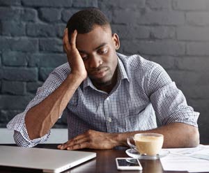 Man at table with his laptop, phone and coffee in front of him, with the side of his face resting in his palm.