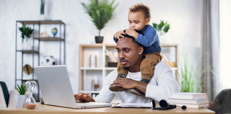 Father sitting in front of laptop at home with young son sitting on his shoulders.