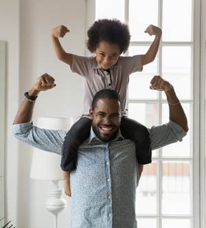 Young boy on man’s shoulders, both raising clenched fists with elbows bent in a “strong-man” pose.