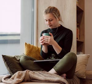 Young woman on couch with laptop, working from home and holding a mug in her hands.