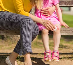 Adult woman and female child seated on a bench, shown from the waist down. Woman is administering first aid to scratch on child’s knee.