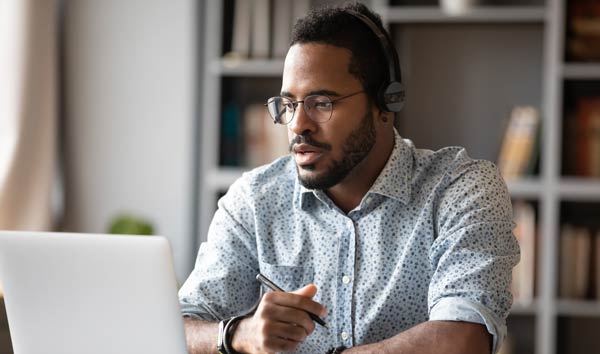 Bearded man with glasses sitting in front of a bookcase, holding a pen and looking at a laptop screen.